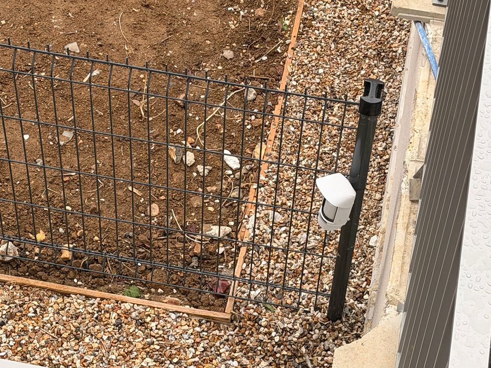 Détecteur extérieur blanc fixé sur un poteau de clôture, au bord d'un terrain en fin de chantier